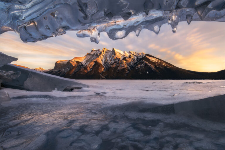 A winter photograph of the Canadian Rockies from Lake Minnewanka in Banff National Park from an ice cave at sunset